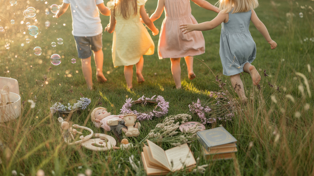 Vier Kinder laufen über eine Wiese. Auf der WIese liegen Bücher, Puppen und gepflückte Blumen