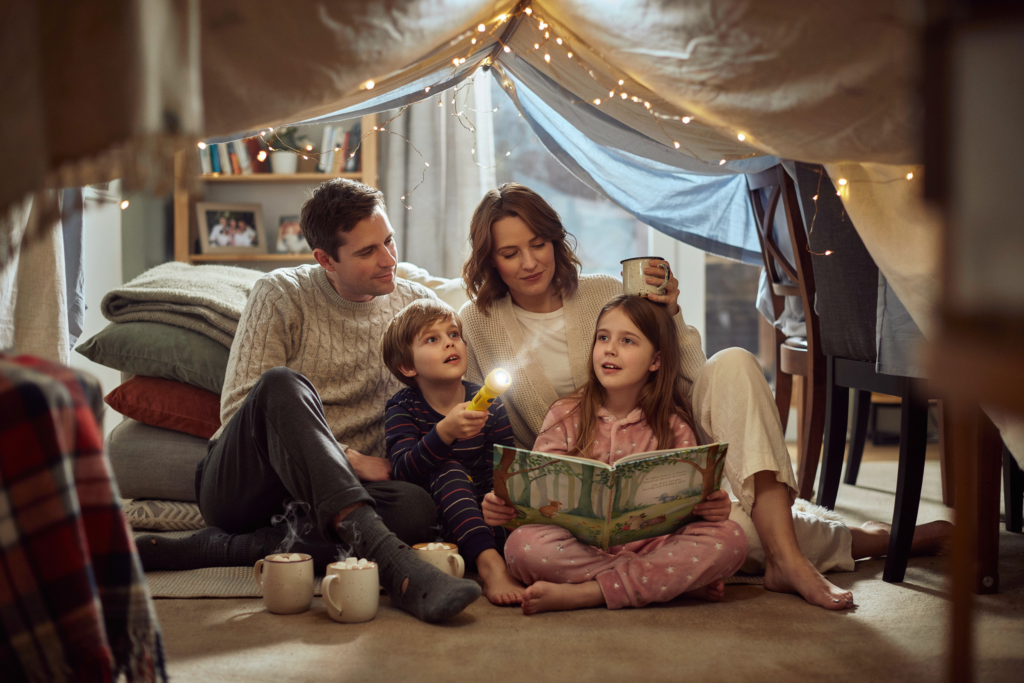 Familie verbringt einen gemütlichen Campingabend im Wohnzimmer und sitzt gemeinsam in einer selbstgebauten Deckenhöhle mit Lichterkette.
