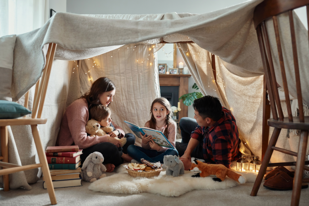 Eltern und Kinder sitzen gemeinsam in einer großen Deckenhöhle mit Lichterkette und genießen einen gemütlichen Familienabend.